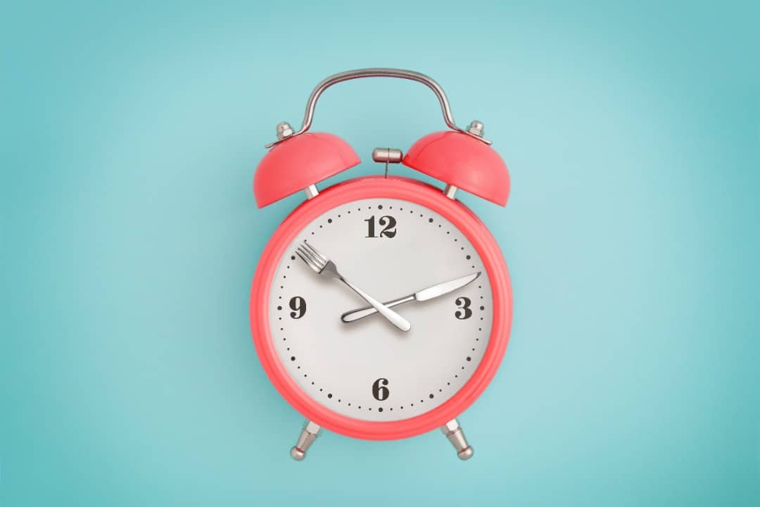 Pink analog alarm clock set on a blue background. The clock hand are a fork and knife.