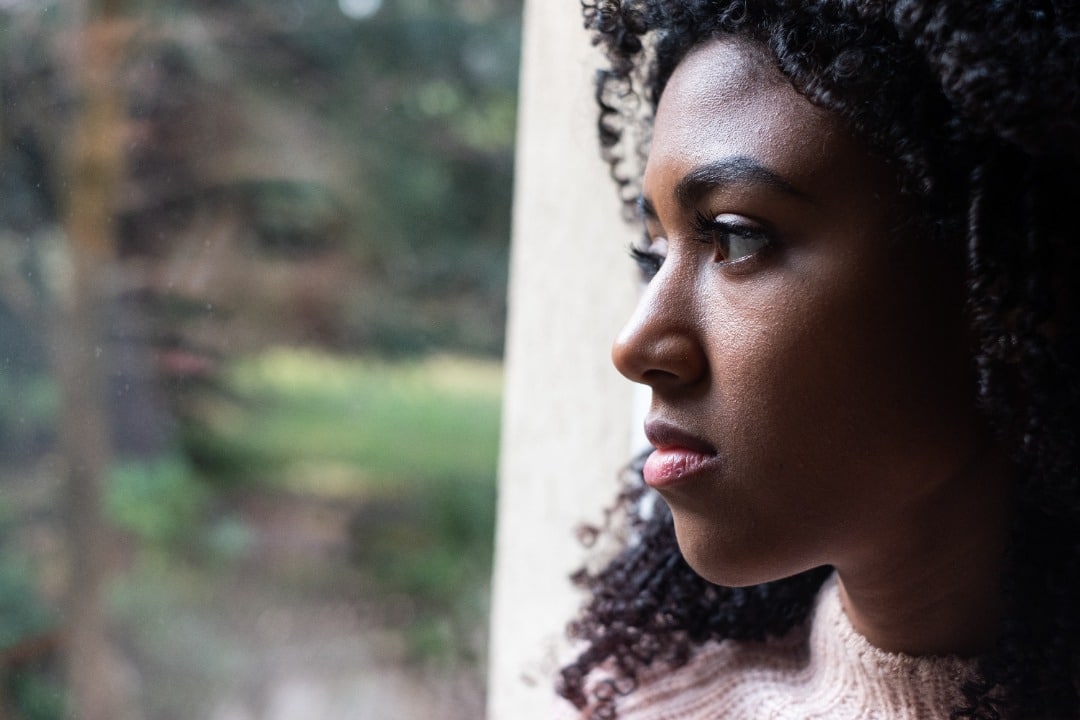 A young black woman in a pink sweater gazes out a window, deep in thought.