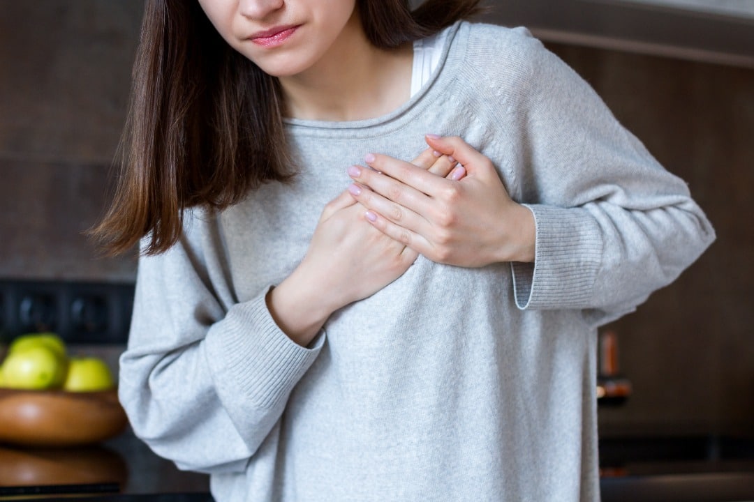 Young woman in gray sweatshirt clutching her chest over her heart in pain.