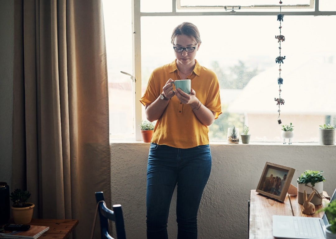 Woman leans against a wall with her back against the window, she's wearing glasses and yellow shirt, and she's holding a cup of coffee.