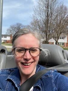 Karen is wearing glasses and smiling at the camera while in a convertible. 
